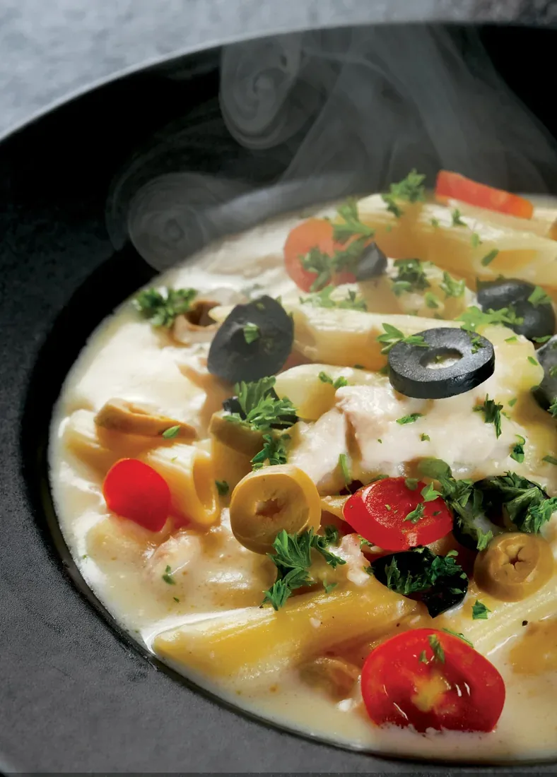 Steaming creamy penne pasta with olives and cherry tomatoes in a black bowl — food photographer in Dubai